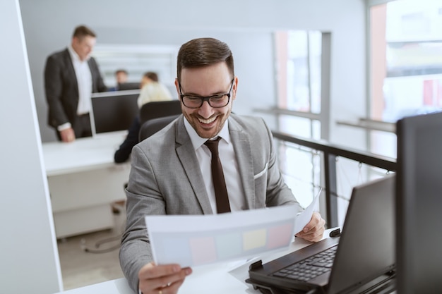 Satisfied Caucasian businessman with eyeglasses and in suit looking at paperwork while sitting at workplace.