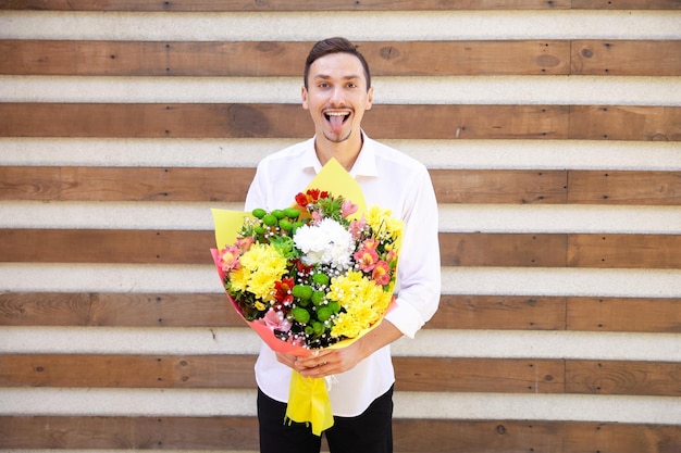 Satisfied Caucasian guy in a white shirt and black jeans showing tongue and holding a big bouquet