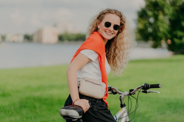 Satisfied curly female bicyclist enjoys spare time travels on bike stops to have little rest wears sunglasses casual summer outfit poses among grass and trees green blurred background