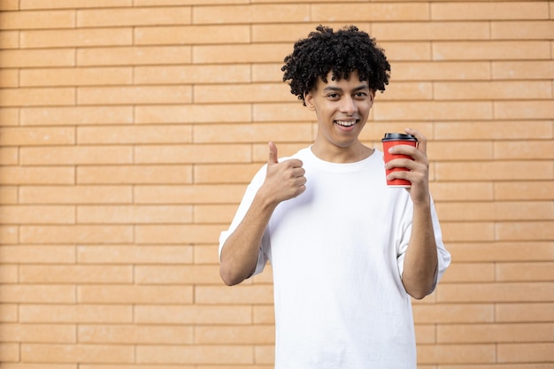 赤い一杯のコーヒーを持って、白いTシャツを着て親指を上げて満足している巻き毛のラテン系の男