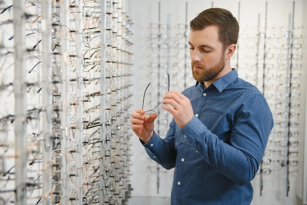 Satisfied Customer View of happy young male client wearing new glasses standing near rack and showcase with eyewear Smiling man trying on spectacles