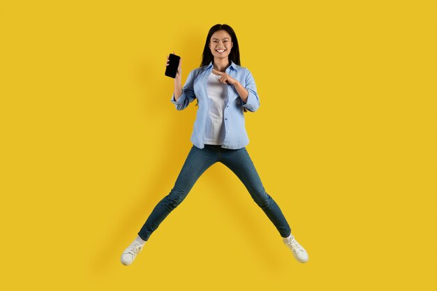 Удовлетворенная симпатичная тысячелетняя азиатская студентка прыгает и замирает в воздухе, указывая пальцем на смартфон с пустым экраном