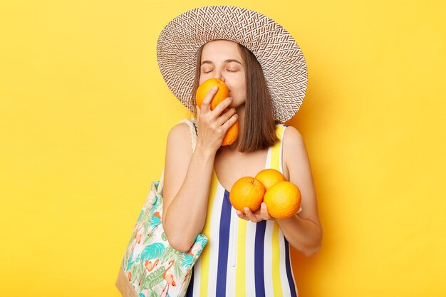 Satisfied delighted woman wearing striped onepiece swimsuit and straw hat posing isolated over yellow background standing smelling oraganic oranges resting on tropical resort