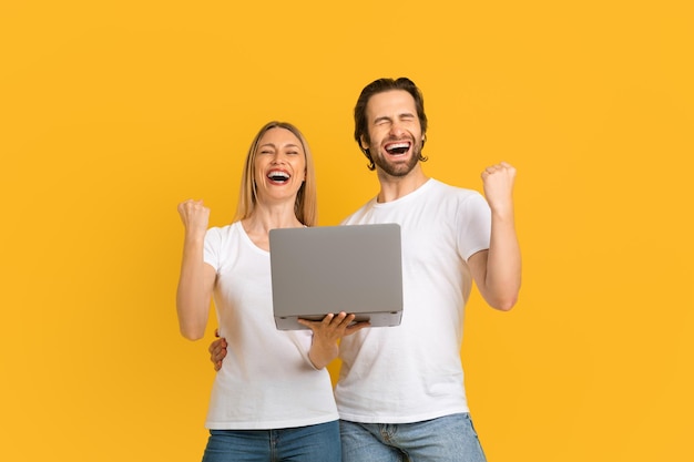 Satisfied excited millennial european couple in white tshirts rejoice in victory and hold laptop