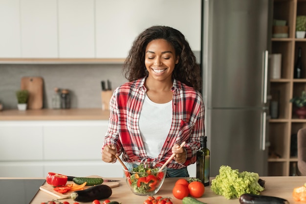 Satisfied glad millennial black female preparing salad at table with organic vegetables has fun at spare time