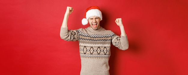 Удовлетворенный красивый мужчина, чувствуя себя удачливым и счастливым, крича от радости и качая кулаками, празднуя победу или победу, получает потрясающий рождественский подарок, стоя в шапке Санты на красном фоне.