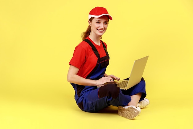Удовлетворенная практичная женщина сидит на полу со скрещенными ногами и работает на ноутбуке, глядя в камеру