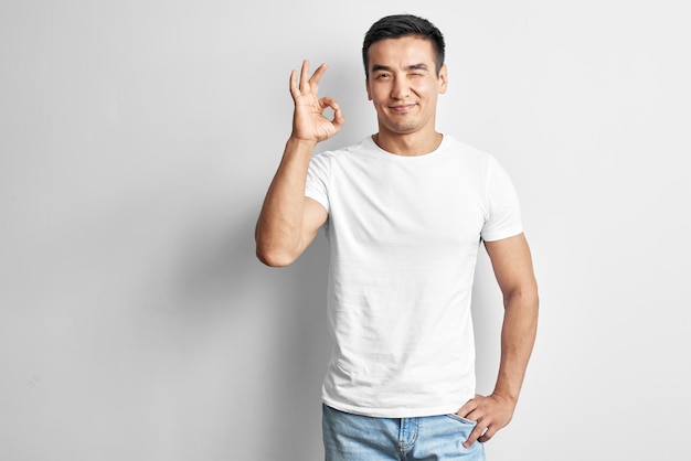 Satisfied Kazakh young man dressed in casual shows the sign "it's OK" on white background with copy space. Joyful successful Asian with pleased facial expression in studio Isolated