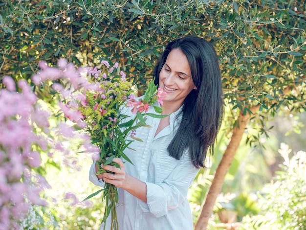  셔츠를 입고 긴 검은 머리카락을 가진 만족스러운 여인이 빛에 꽃이 피는 정원에서 서 있습니다.