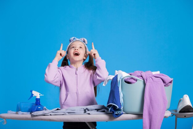 The satisfied little girl smiled and found a solution to problems in ironing the washed clothes Child points two fingers upward with delight portrait on blue background advertising space