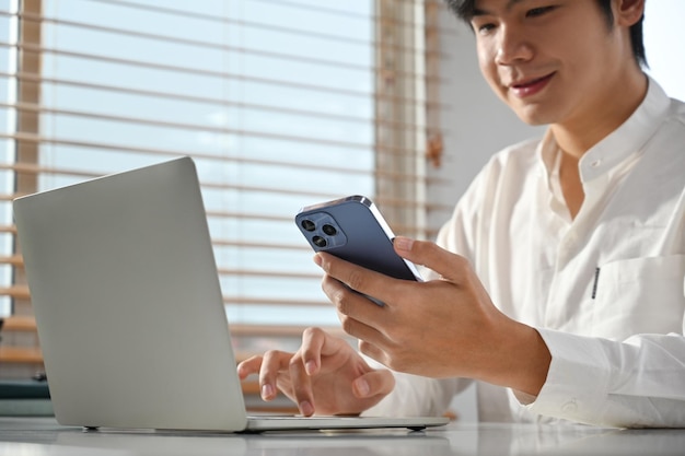 Satisfied male entrepreneur chatting with client online on smart phone and using laptop at modern workplace