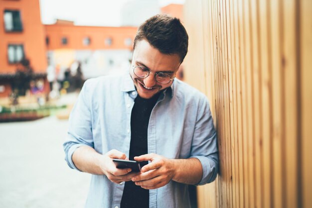 Satisfied man in casual outfit smiling while checking photo o smartphone at city street