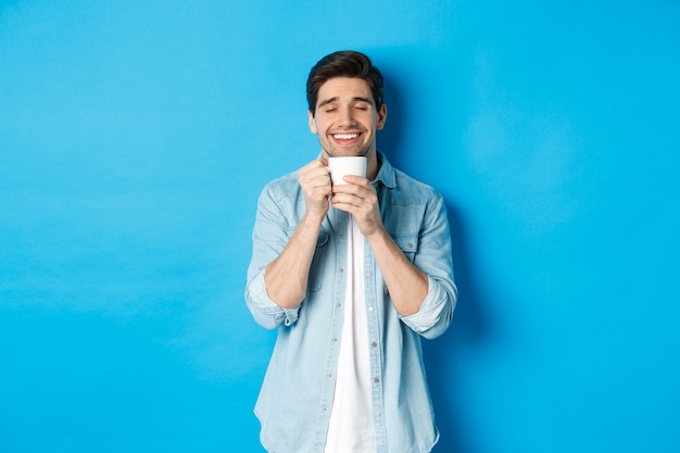 Satisfied man enjoying cup of tea or coffee, holding mug with pleased smile, standing against blue wall