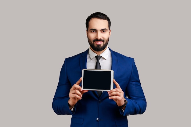 Satisfied man showing tablet empty screen for adv, has happy facial expression, looking at camera with toothy smile, wearing official style suit. Indoor studio shot isolated on gray background.