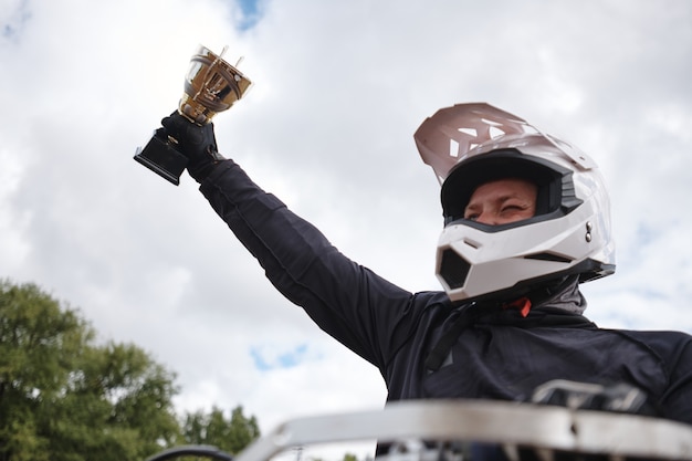 Satisfied man in white helmet raising hand with motorcycle competition cup after winning competition