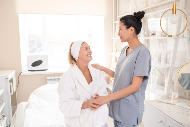 Satisfied mature beauty salon guest in robe sitting on procedure table and holding hand of Asian cosmetologist while thanking her after procedure
