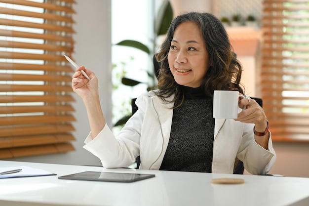 La donna soddisfatta di affari maturi che tiene una tazza di caffè e guarda attraverso la finestra gode di un'atmosfera pacifica e senza stress