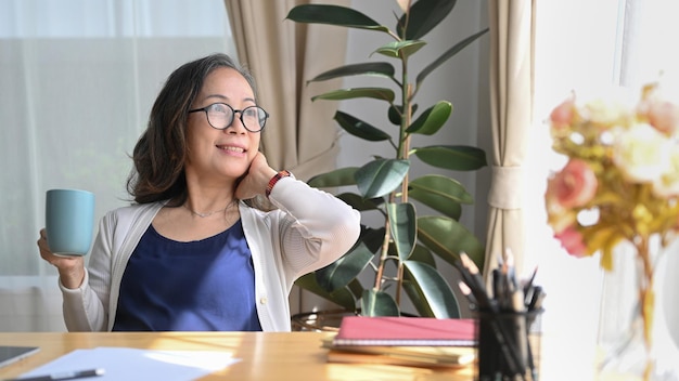 Satisfied middle aged woman holding coffee cup and looking through the window enjoy stress free peaceful mood