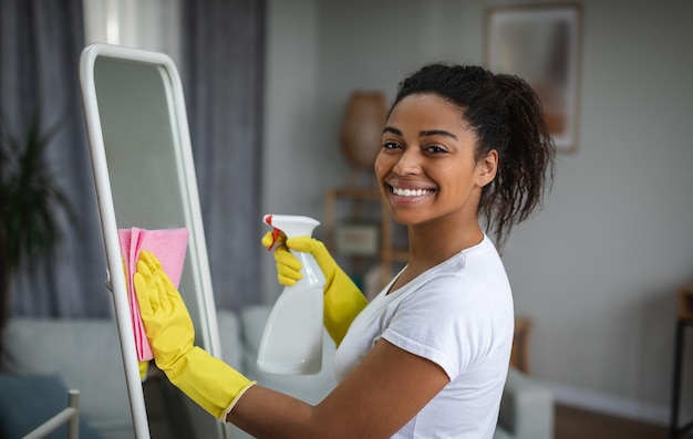 Satisfied millennial african american woman housewife in rubber gloves washing mirror with spray and microfiber rag