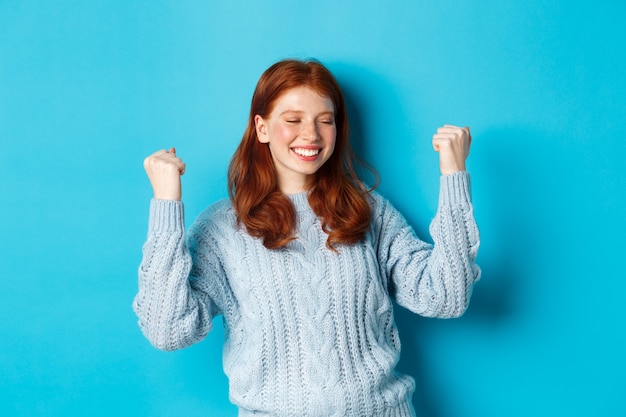 La ragazza rossa soddisfatta raggiunge l'obiettivo e festeggia, facendo il gesto della pompa del pugno e sorridendo con gioia, trionfando della vittoria, in piedi su sfondo blu.