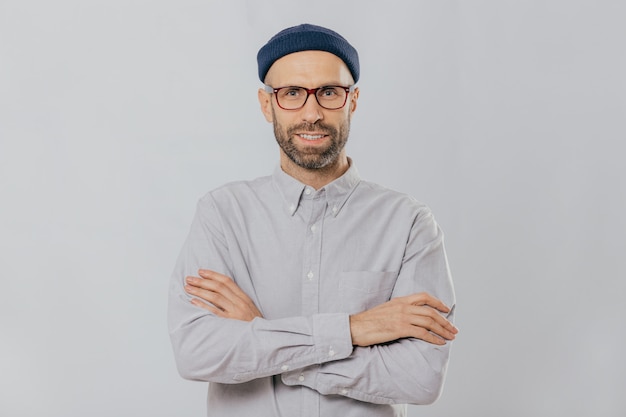 Satisfied self confident male designer wears stylish headgear, dressed in white shirt, keeps arms folded