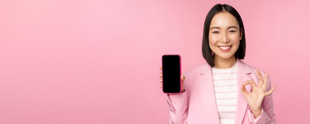 Satisfied smiling asian businesswoman recommending mobile phone app website company on smartphone showing screen and okay sign pink background