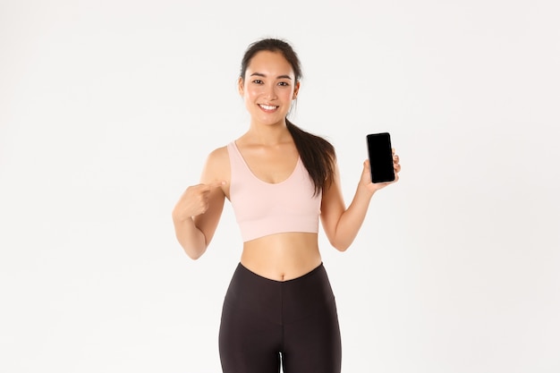 Ragazza asiatica sorridente soddisfatta di forma fisica, schermo dello smartphone del dito puntato sportivo attraente.