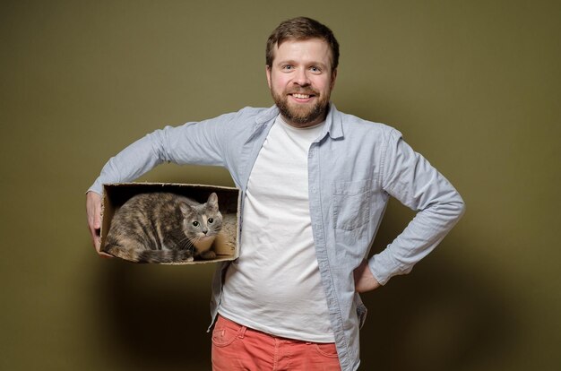 Satisfied smiling bearded man holds cardboard box in which beloved cat sits and looks with interest