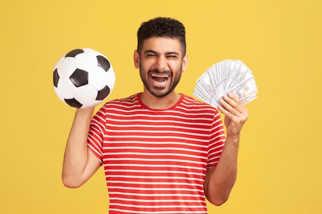 写真 サッカーボールとドル紙幣のファン、スポーツベッティング、大勝利を保持しているストライプのtシャツのひげで満足した笑顔の男。黄色の背景に分離された屋内スタジオショット