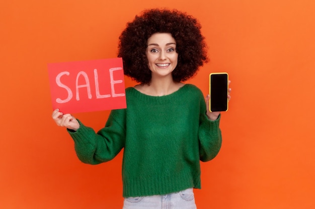 Satisfied smiling woman with Afro hairstyle wearing green casual style sweater holding sale card and smart phone with blank black display Indoor studio shot isolated on orange background