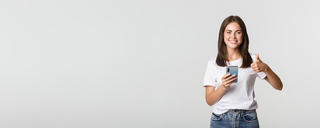 Satisfied smiling young woman showing thumbsup while using smartphone