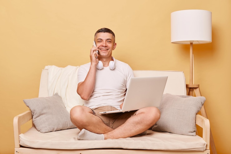 Photo satisfied smilng man wearing white tshirt and shorts sitting on sofa against beige background remote job working on computer talking phone while having break