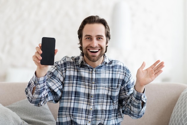 Удовлетворенный удивленный взволнованный молодой европейский мужчина в рубашке жестикулирует телефон шоу с пустым экраном
