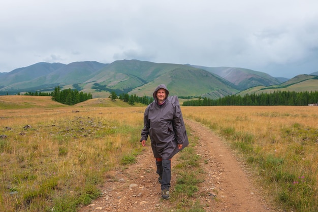 Satisfied tourist returns from mountains in overcast Happy man in raincoat walks through hills and forest in bad weather Traveler goes towards adventure Hiker and mountain range under cloudy sky