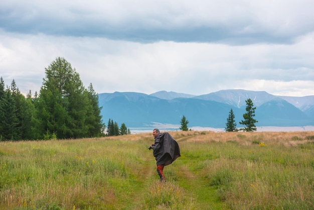 Satisfied tourist with camera in mountains in overcast Happy man in raincoat walks through hills and forest in bad weather Traveler goes towards adventure Hiker and mountain range under cloudy sky