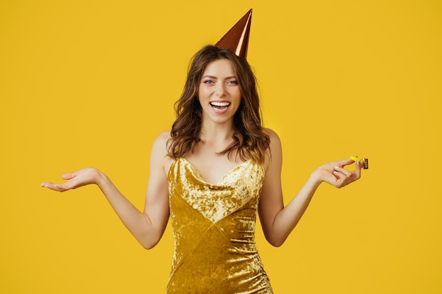 Satisfied woman in elegant dress and birthday cap holding party horn looking and smiling at camera