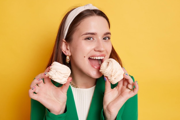 Satisfied woman wearing green jacket posing isolated over yellow background holding biting marshmallow breaking diet eating sugary desserts