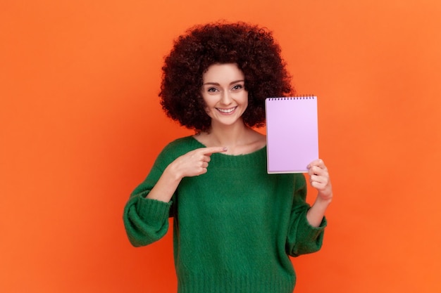 Satisfied woman with Afro hairstyle in green casual style sweater standing showing paper organizer, pointing at notebook, look at camera with smile. Indoor studio shot isolated on orange background.