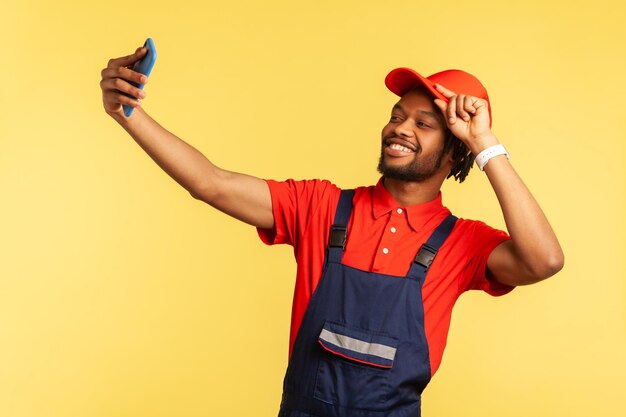 Satisfied worker in overalls taking selfie or talking on video call, looking playfully at device camera to assure client about quality online order. Indoor studio shot isolated on yellow background.