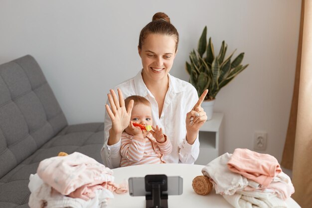 Satisfied young adult woman posing with infant daughter in front of tripod with phone, filming video for her vlog or broadcasting livestream, showing age of kid with fingers.