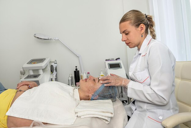 satisfied young beautiful woman uses the services of a professional cosmetologist in a beauty salon or spa beautician applied a white mask on the client's face with a brush View from above