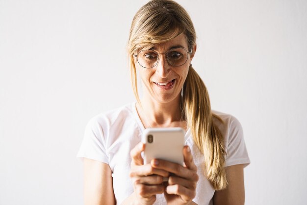 Satisfied young blond girl types text message on cell phone\
enjoys online communication types feedback wears white shirt\
technology concept image