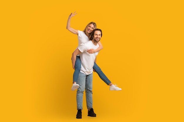 Satisfied young european guy with stubble in white tshirt hold lady on his back female shows peace