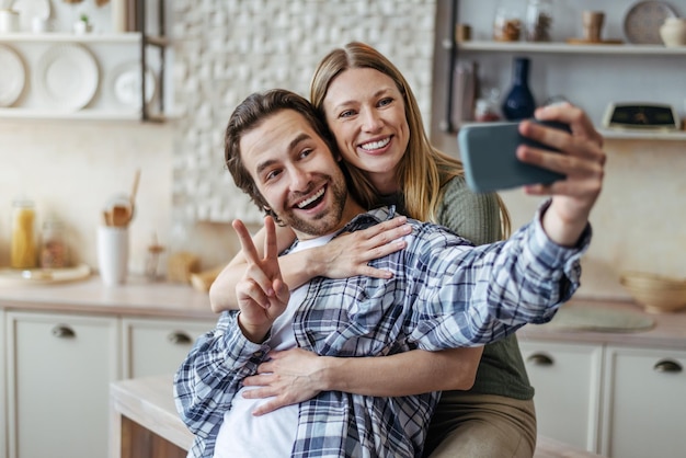 Satisfied young european lady hugging male with stubble guy shows peace sign makes selfie on phone