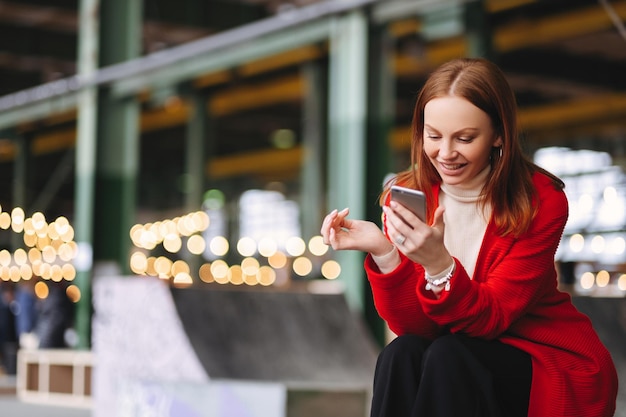 Satisfied young European woman in red coat holds cell phone reads notification connected to wireless internet has spare time poses outdoor waits for someone enjoys online communication