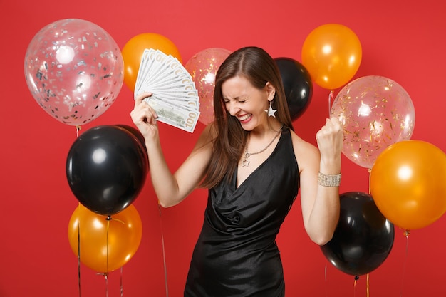 Satisfied young girl in black dress holding bundle lots of dollars cash money doing winner gesture on red background air balloons. Valentine's Day Happy New Year birthday mockup holiday party concept.