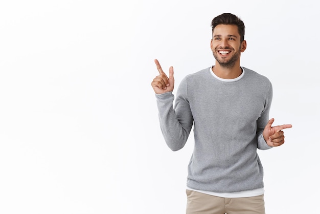 Satisfied young handsome man with bristle wander around shopping mall stalls surrounded good choices explore interesting place pointing sideways decide where travel during winter holiday smiling