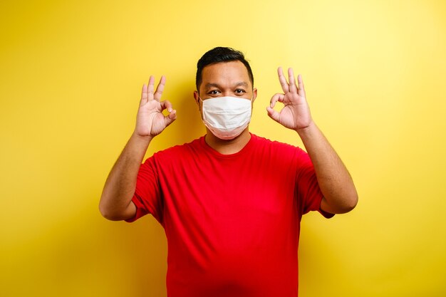 Satisfied young man in medical mask showing make okay sign against yellow background