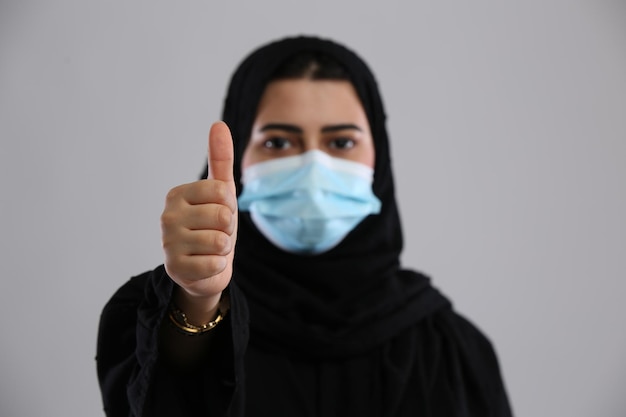 Photo saudi arabia woman in abaya wearing mask standing with thumbs up sign thumb in focus