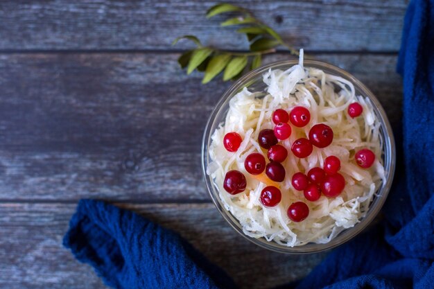 Sauerkraut with cranberries in glass bowl with blue towel on wooden background top view